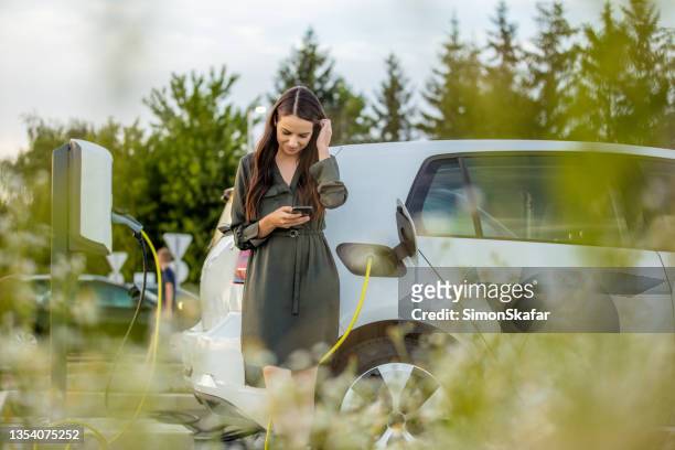 woman using mobile phone while waiting for electric car to charge in the parking lot - charging stockfoto's en -beelden