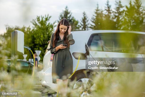 woman using mobile phone while waiting for electric car to charge in the parking lot - electric car stock pictures, royalty-free photos & images