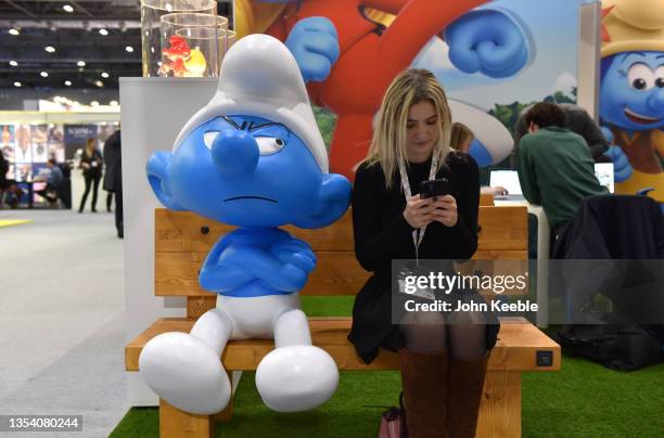 Woman looks at her phone next to a Grouchy Smurf character at the Smurfs stand during Brand Licensing Europe at ExCel on November 18, 2021 in London,...