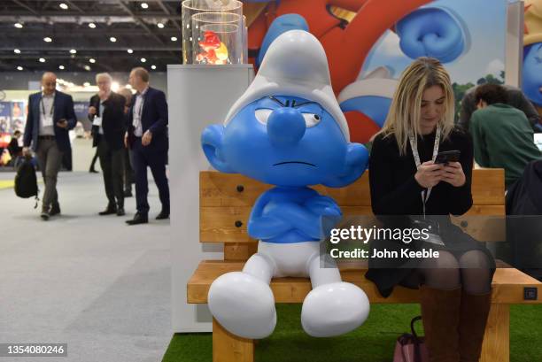 Woman looks at her phone next to a Grouchy Smurf character at the Smurfs stand during Brand Licensing Europe at ExCel on November 18, 2021 in London,...