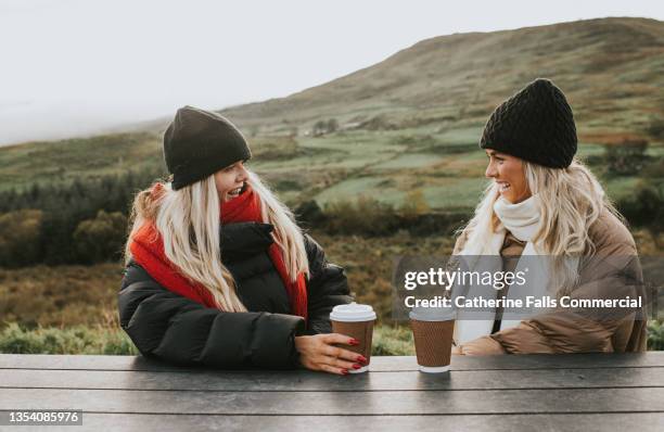 two beautiful young woman in warm clothing, enjoy a hot drink at an outdoor picnic table - sitting bench stock pictures, royalty-free photos & images