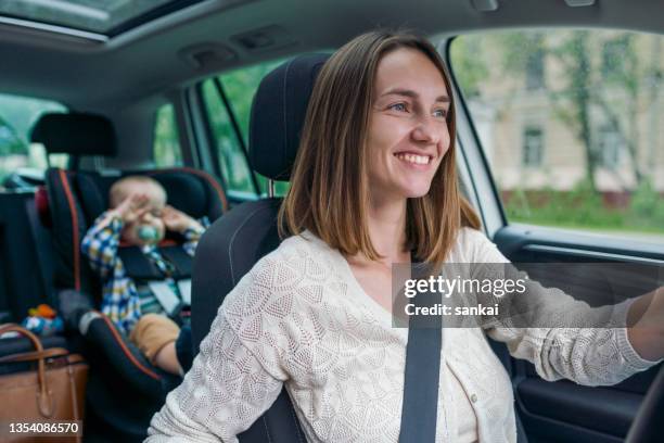 hermosa mujer sonriente conduciendo un automóvil con un pequeño bebé en el asiento trasero - woman driving fotografías e imágenes de stock