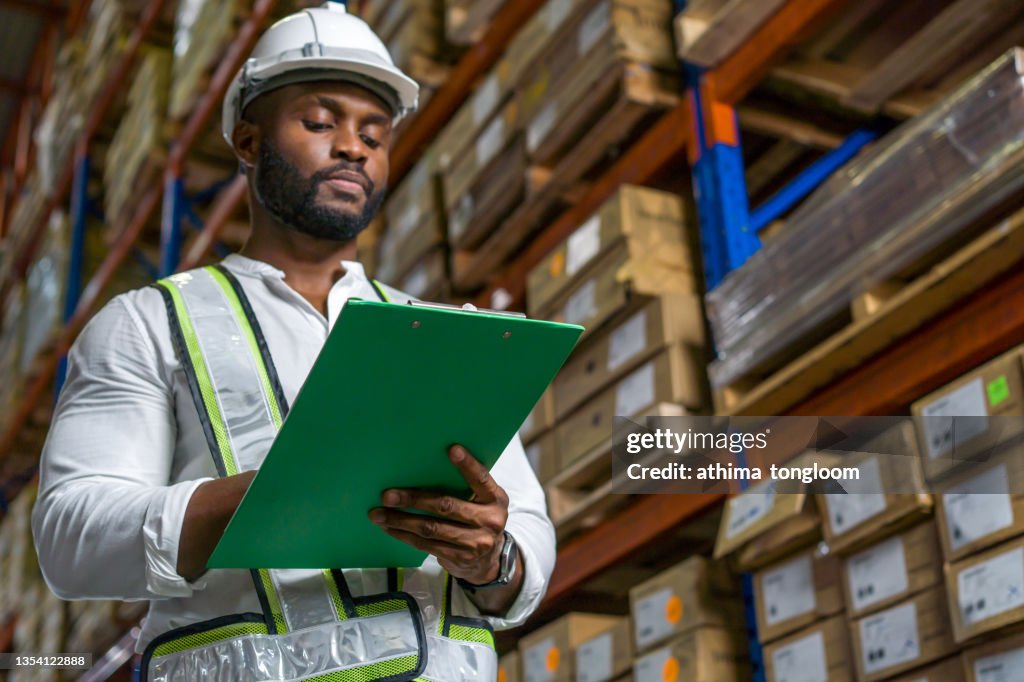 Smart engineer man wearing doing stock tick check and cardboard stock product management in factory warehouse background