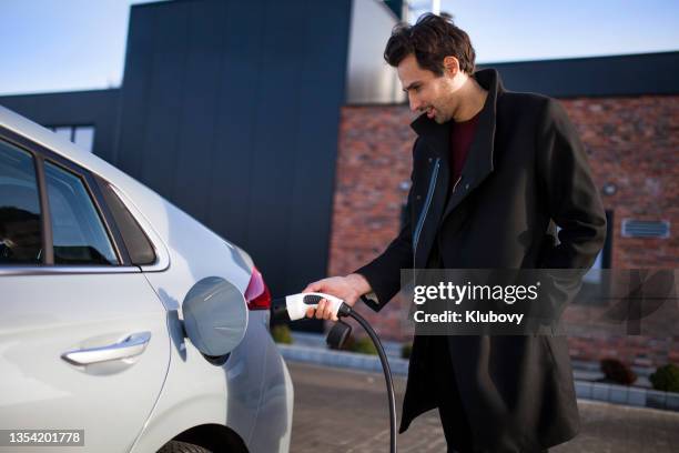 junger mann auf einem parkplatz, der sein elektroauto auflädt - elektroauto mensch stock-fotos und bilder