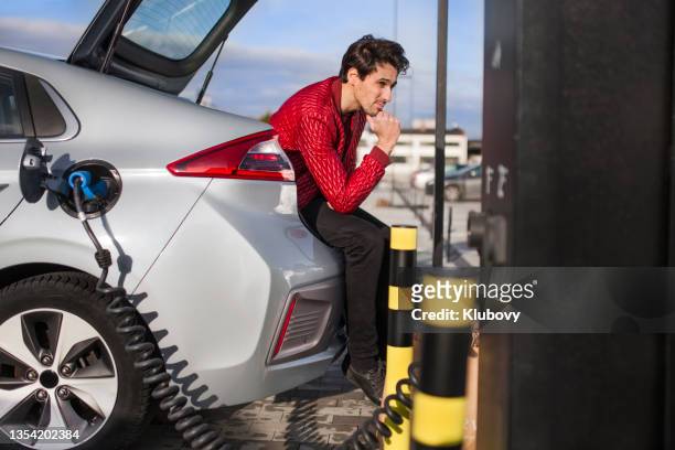 young man on a parking lot, charging his electric car - red car wire 個照片及圖片檔