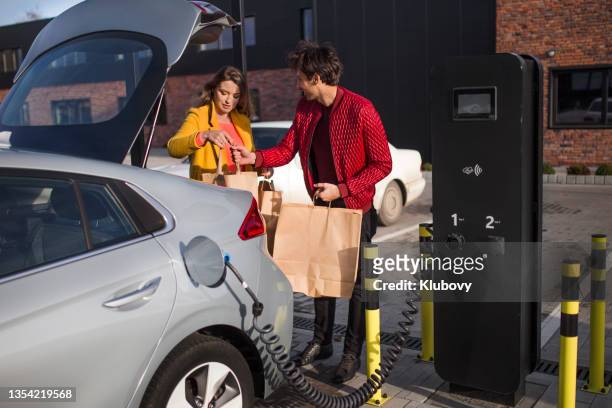 young couple on a store parking lot, charging their electric car - red car wire 個照片及圖片檔