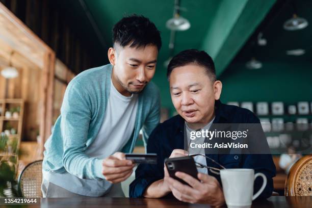 joyful asian senior father and son shopping online with mobile app on smartphone together and making payment with credit card online at home - old asian man stockfoto's en -beelden