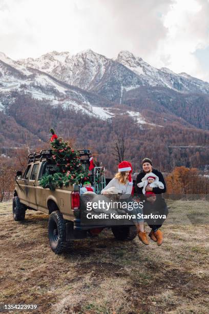 young european and young korean and their newborn daughter are sitting in a decorated christmas pickup with a tree and a white dog golden retriever against the backdrop of snow-capped mountains - funny people stock pictures, royalty-free photos & images