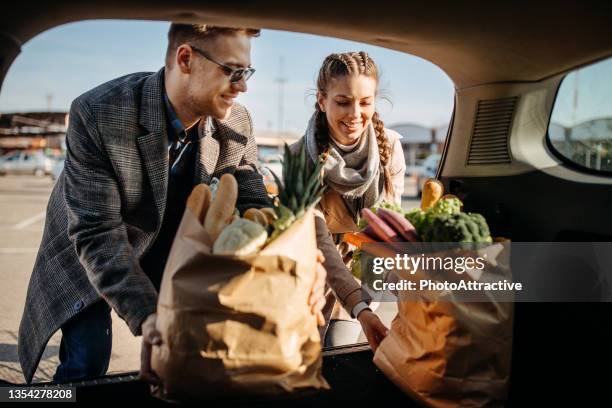 happy young couple putting groceries in a car at parking lot - achterbak-van-auto stockfoto's en -beelden
