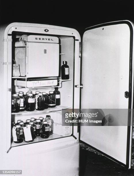 The door of a refrigerator is open showing two shelves of bottles of blood. A sign on the tray below the freezer reads: '94th Evac. Hosp.'.