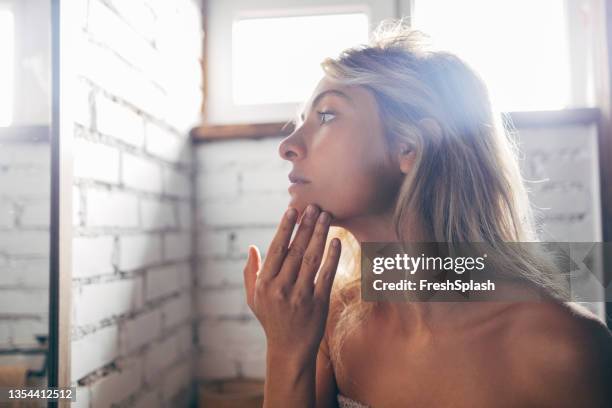 ritratto di una bella donna bionda in piedi davanti allo specchio del bagno e applicando la crema da giorno per il viso - corpo normale foto e immagini stock