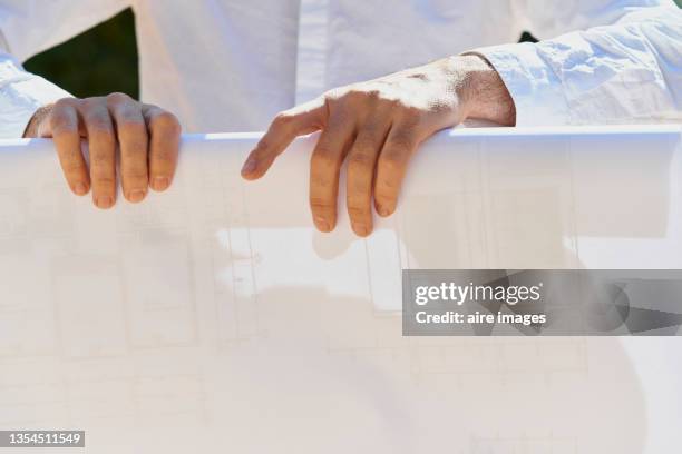 close-up of front view of cropped hands of a male contractor rolling up blueprints of a project in the construction site out of building during sunny day - architekt mit plan frontal stock-fotos und bilder