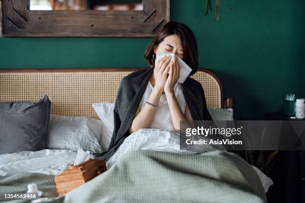 joven asiática sentada en la cama y sonándose la nariz con pañuelos de papel mientras sufría de un resfriado, con un frasco de medicamentos y un vaso de agua en la mesa auxiliar - sonarse fotografías e imágenes de stock