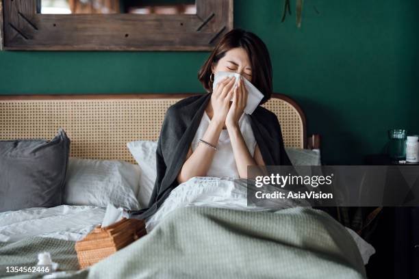 jeune femme asiatique assise sur son lit et se mouchant avec un mouchoir en papier tout en souffrant d’un rhume, avec un flacon de médicament et un verre d’eau sur la table d’appoint - virus grippal photos et images de collection