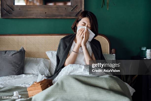 giovane donna asiatica seduta sul letto e soffiarsi il naso con un fazzoletto mentre soffre di raffreddore, con bottiglia di medicina e un bicchiere d'acqua sul tavolino - virus grippe foto e immagini stock