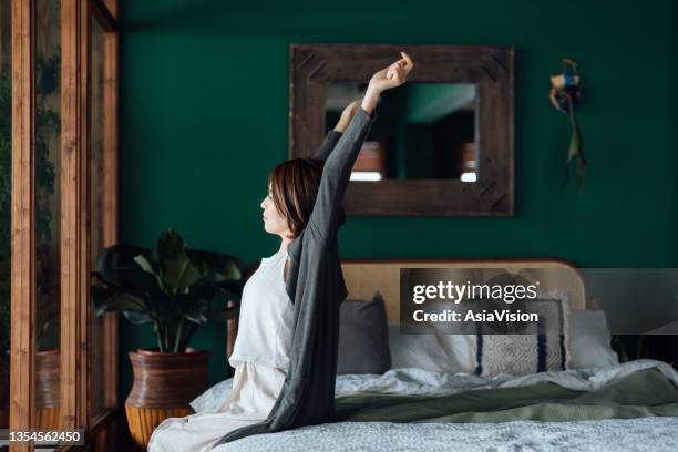 side profile of young asian woman sitting in bed, stretching arms after waking up in the morning against sunlight. let's get the day started - woman waking up happy stock pictures, royalty-free photos & images