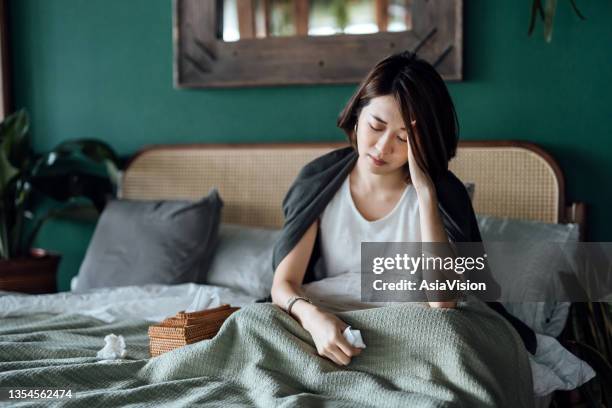 young asian woman feeling sick and suffering from a headache, massaging forehand to relieve the pain, sitting on the bed and taking a rest at home - chronic illness stock pictures, royalty-free photos & images