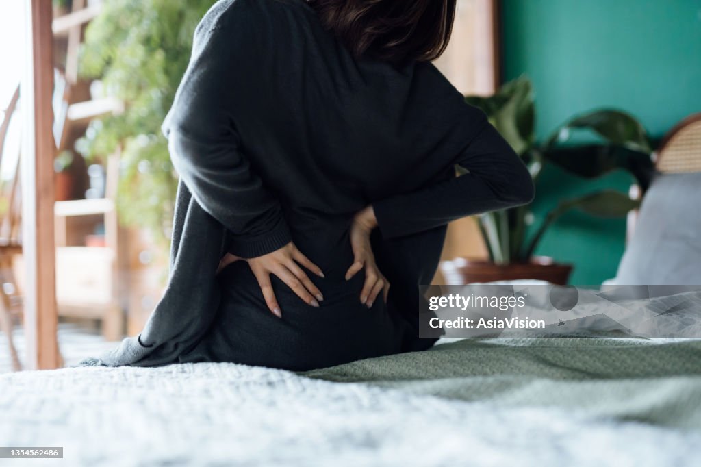 Rear view of woman suffering from backache, massaging aching muscles while sitting on bed