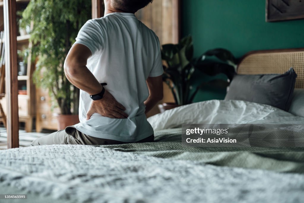 Rear view of senior Asian man suffering from backache, massaging aching muscles while sitting on bed. Elderly and health issues concept