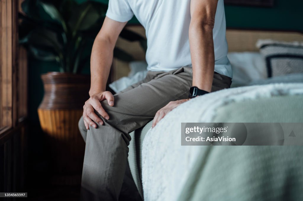Close up of senior man holding his knee in discomfort, suffering from knee pain while sitting on bed at home. Elderly and health issues concept