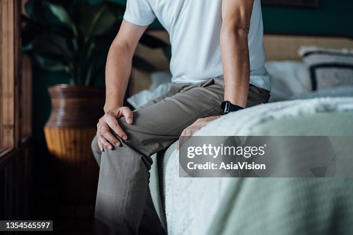 Close up of senior man holding his knee in discomfort, suffering from knee pain while sitting on bed at home. Elderly and health issues concept Close up of senior man holding his knee in discomfort, suffering from knee pain while sitting on bed at home. Elderly and health issues concept