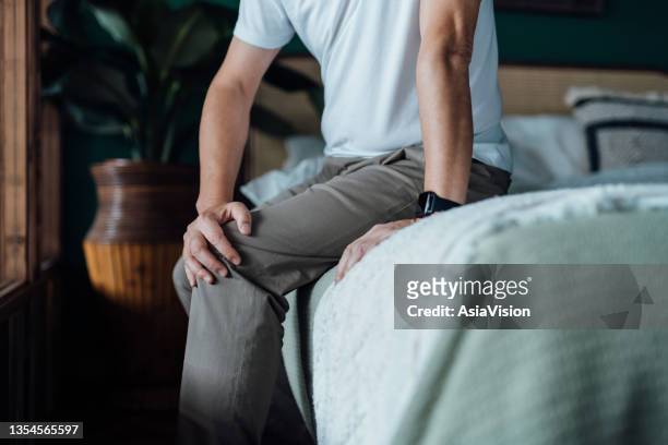 close up of senior man holding his knee in discomfort, suffering from knee pain while sitting on bed at home. elderly and health issues concept - rest cure stock pictures, royalty-free photos & images