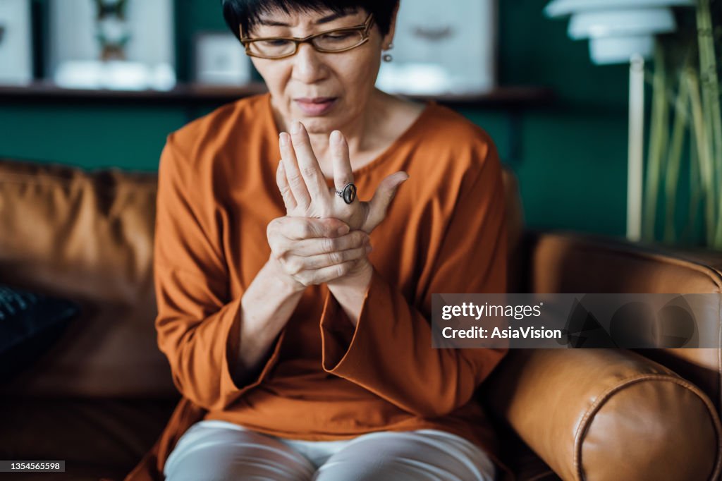 Mujer asiática mayor frotándose las manos con incomodidad, sufriendo de artritis en la mano mientras está sentada en el sofá de su casa. Concepto de ancianos y problemas de salud