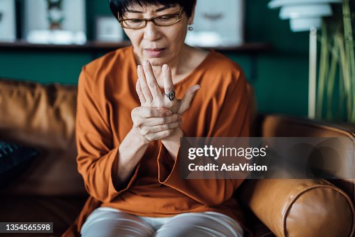 Senior Asian woman rubbing her hands in discomfort, suffering from arthritis in her hand while sitting on sofa at home. Elderly and health issues concept Senior Asian woman rubbing her hands in discomfort, suffering from arthritis in her hand while sitting on sofa at home. Elderly and health issues concept