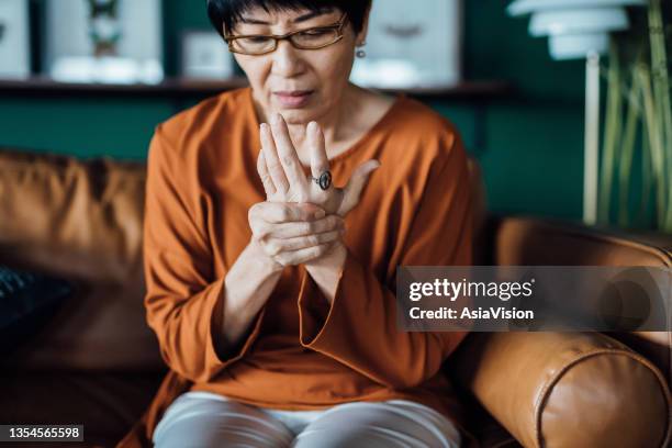 senior asian woman rubbing her hands in discomfort, suffering from arthritis in her hand while sitting on sofa at home. elderly and health issues concept - inflamação imagens e fotografias de stock