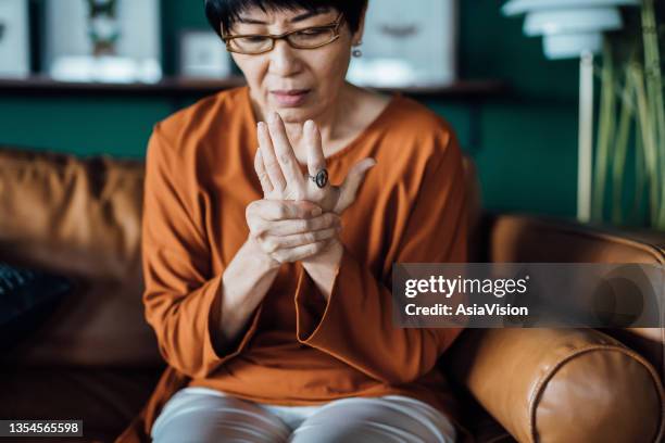 senior asian woman rubbing her hands in discomfort, suffering from arthritis in her hand while sitting on sofa at home. elderly and health issues concept - tender imagens e fotografias de stock