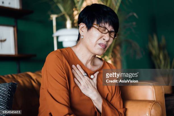 senior asian woman with eyes closed holding her chest in discomfort, suffering from chest pain while sitting on sofa at home. elderly and health issues concept - heart attack stockfoto's en -beelden