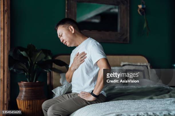 senior asian man with eyes closed holding his chest in discomfort, suffering from chest pain while sitting on bed at home. elderly and health issues concept - stroke illness stockfoto's en -beelden