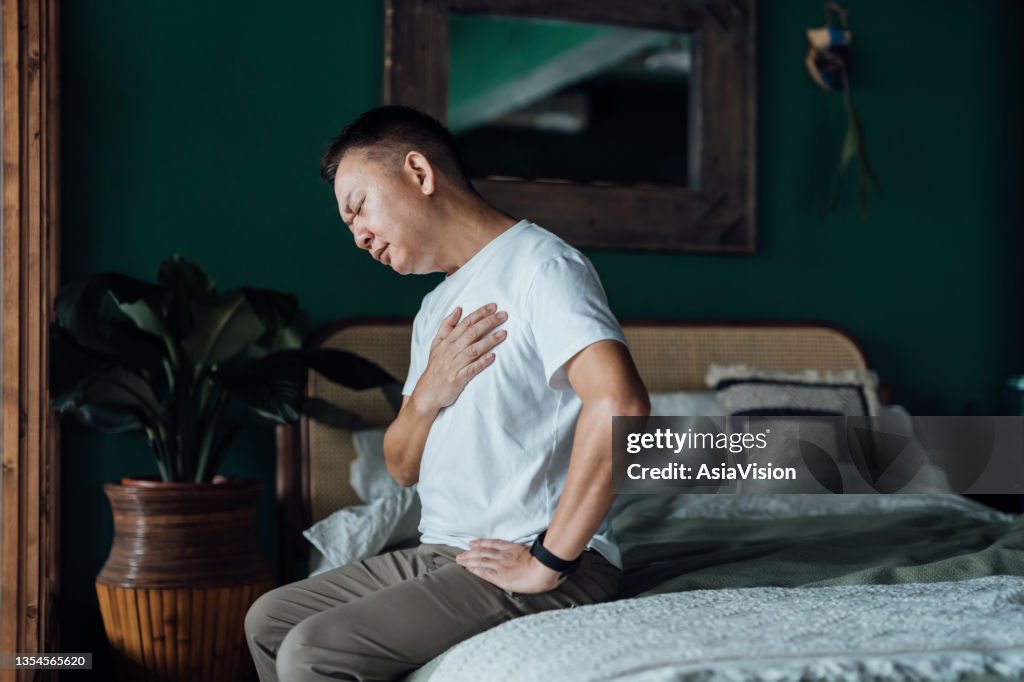 Senior Asian man with eyes closed holding his chest in discomfort, suffering from chest pain while sitting on bed at home. Elderly and health issues concept