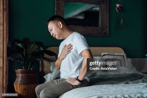 senior asian man with eyes closed holding his chest in discomfort, suffering from chest pain while sitting on bed at home. elderly and health issues concept - cardiovascular system 個照片及圖片檔