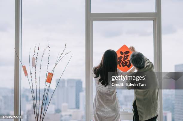 vista posteriore capodanno cinese donna anziana e sua figlia che decora il soggiorno della casa con decorazione calligrafica cinese sulla finestra che si prepara per la riunione di famiglia - chinese new year foto e immagini stock