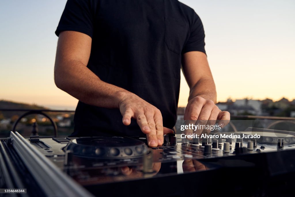 Unrecognizable male DJ hands playing music in a session with a mixer at a sunset party on a terrace.