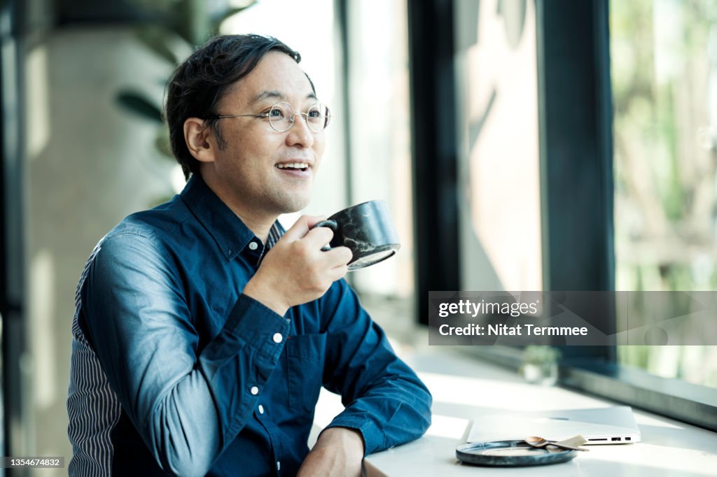 Side of a Confident Japanese businessman holding a coffee mug and looking away by a window in a cafe.