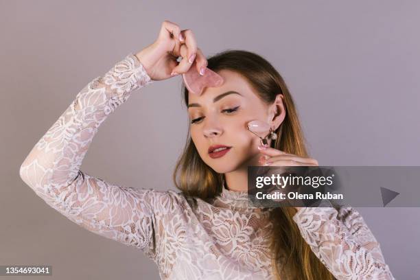 young woman with gua sha scraper and roller in light dress does a facial massage. - jadesteine stock-fotos und bilder