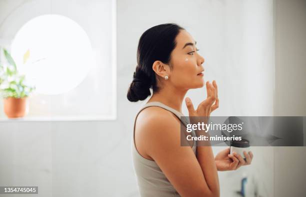 morning routine: attractive asian woman applying face cream in her home - applying makeup stockfoto's en -beelden