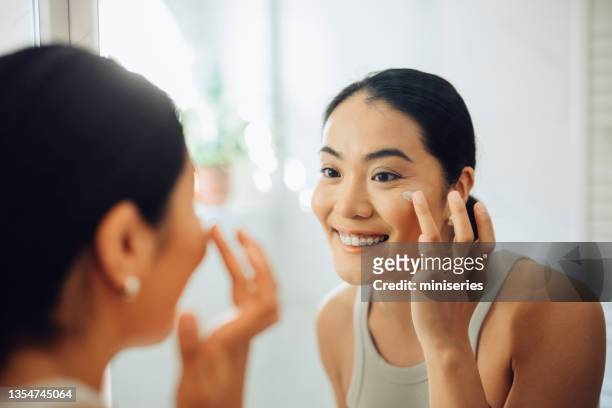 routine du matin: une femme asiatique attrayante applique de la crème pour le visage chez elle - applying stock photos et images de collection