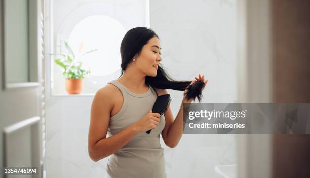 朝のルーチン:彼女のバスルームで彼女の髪をブラッシング美しい幸せなアジアの女性 - hair products ストックフォトと画像
