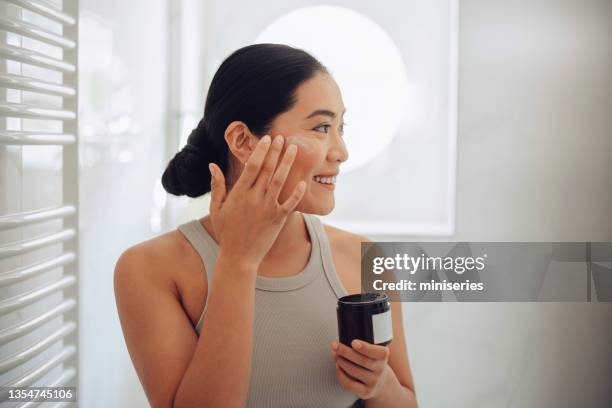 morning routine: attractive asian woman applying face cream in her home - face cream stock pictures, royalty-free photos & images