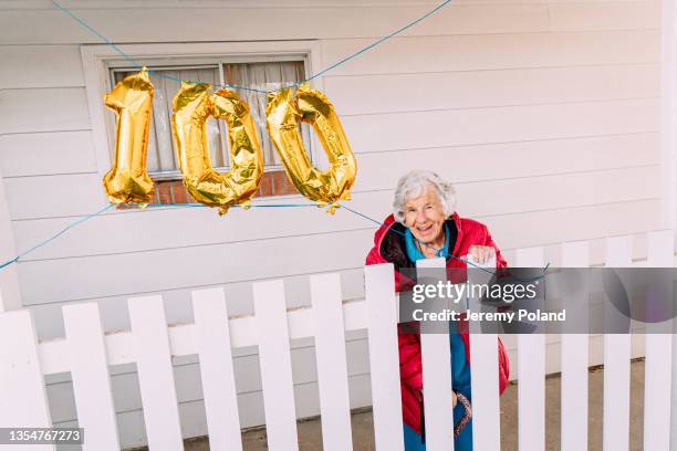 wide angle shot of a cheerful elderly senior caucasian woman leaning proudly on a fence and celebrating her 100th birthday with golden balloons - centenário imagens e fotografias de stock