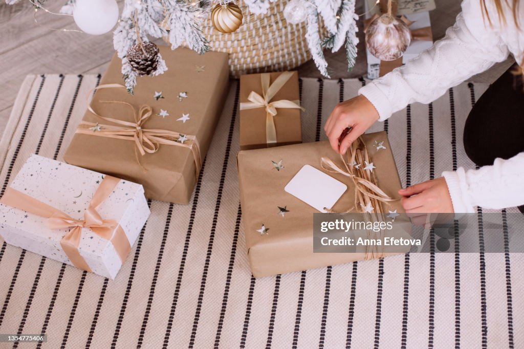 Woman is unpacking gift box with white tag near white and beige paper gift boxes on carpet near decorated Christmas tree in wicker basket. New Year celebration concept. Front view