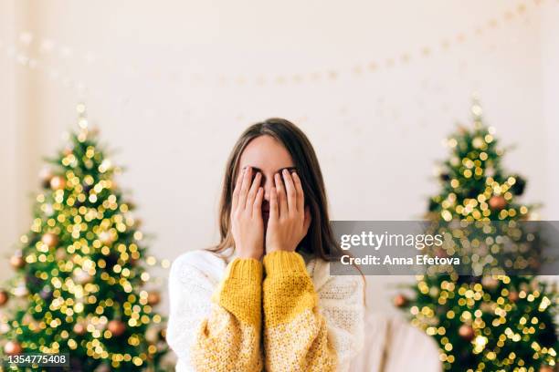 woman in white knitted sweater with yellow sleeves is covering her face with hands on background with two defocused decorated christmas tree with lights. new year celebration concept. front view - double facepalm stock pictures, royalty-free photos & images
