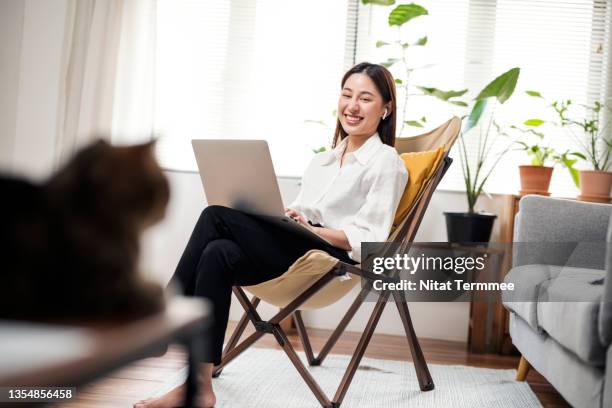 smiling asian women sitting on camping chair working on a laptop while looking to her cat during taking a brake on business meeting at her apartment. cat lover, relaxing during working at home. - cat laptop stock pictures, royalty-free photos & images