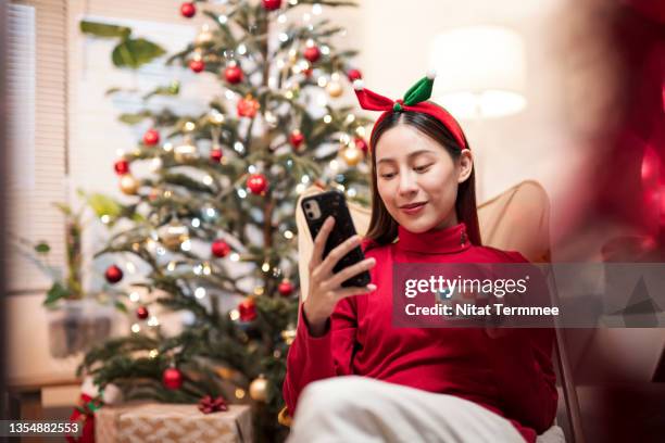 young woman using smartphone during christmas day while sitting on a folding chair in front of christmas tree at her apartment. digital activity increases during new year’s holiday. - years of the conran shop stockfoto's en -beelden