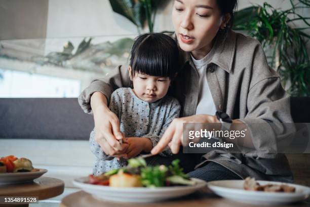 Young Asian mother and lovely little daughter having lunch in cafe, they are cutting the food served on table together, enjoying a happy meal together. Family, food and lifestyle concept