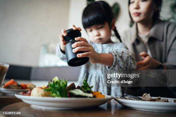 young asian mother and lovely little daughter enjoying lunch in cafe, daughter adding salt into the food served on table, sharing healthy food together. family, food and lifestyle concept - gesalzenes stock-fotos und bilder