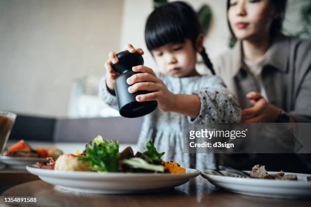 young asian mother and lovely little daughter enjoying lunch in cafe, daughter adding salt into the food served on table, sharing healthy food together. family, food and lifestyle concept - salt shaker ストックフォトと画像
